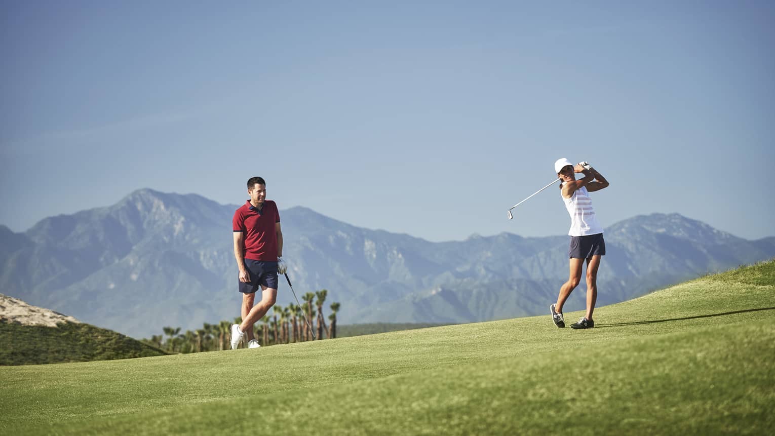 Woman swings golf club as man watches on green course, mountains in background
