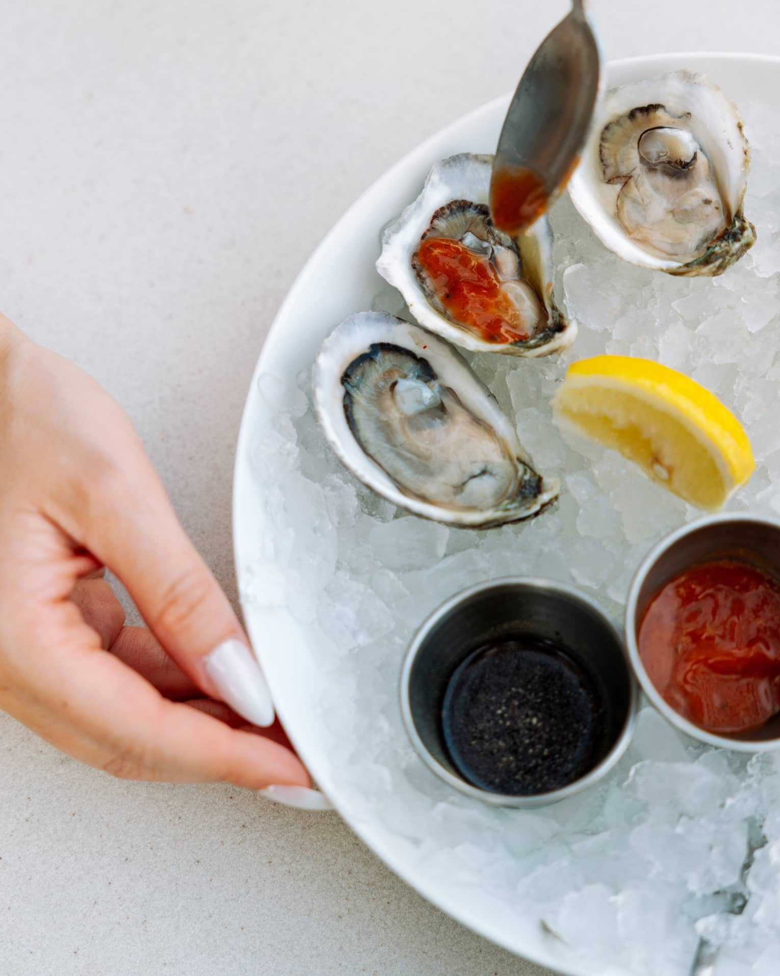 A manicured hand by a bowl of shucked oysters drizzled with red sauce, a lemon wedge and sauce cups on a bed of crushed ice.