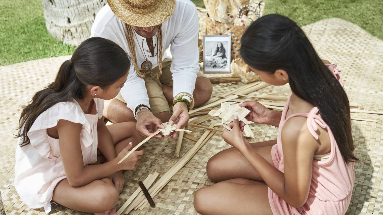 Adult helps two young girls learn how to weave as they sit on a woven blanket
