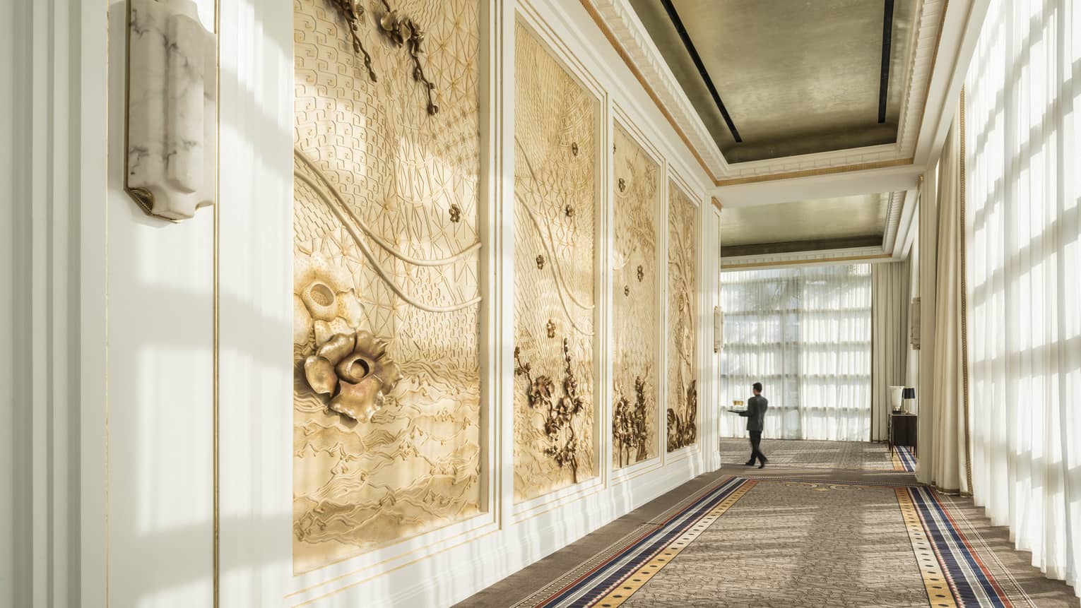 Hotel staff carries tray at end of long hallway past decorative paneled walls