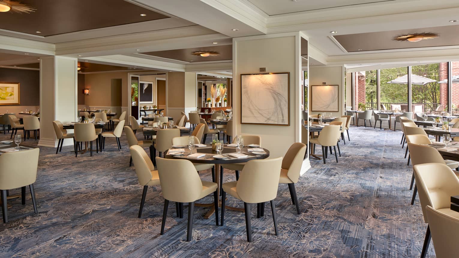 A well lit restaurant with beige chairs and round tables and large open windows.