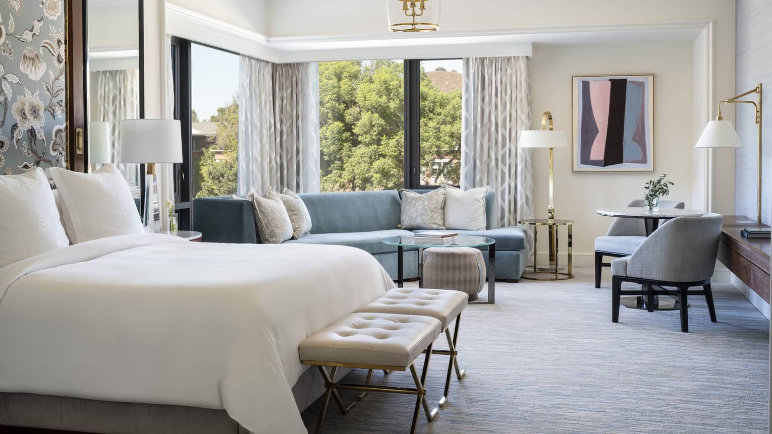 Sunlit corner studio suite with blue furnishings, carpet, drapes, view of trees.