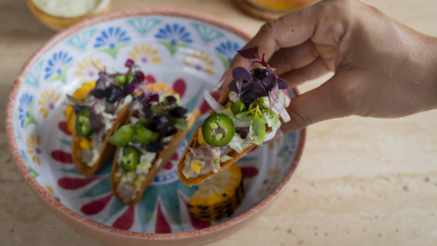 A hand holds a hard-shell taco topped with jalapenos, onions and green-and-purple garnish with two more tacos in a colourful, patterned bowl below