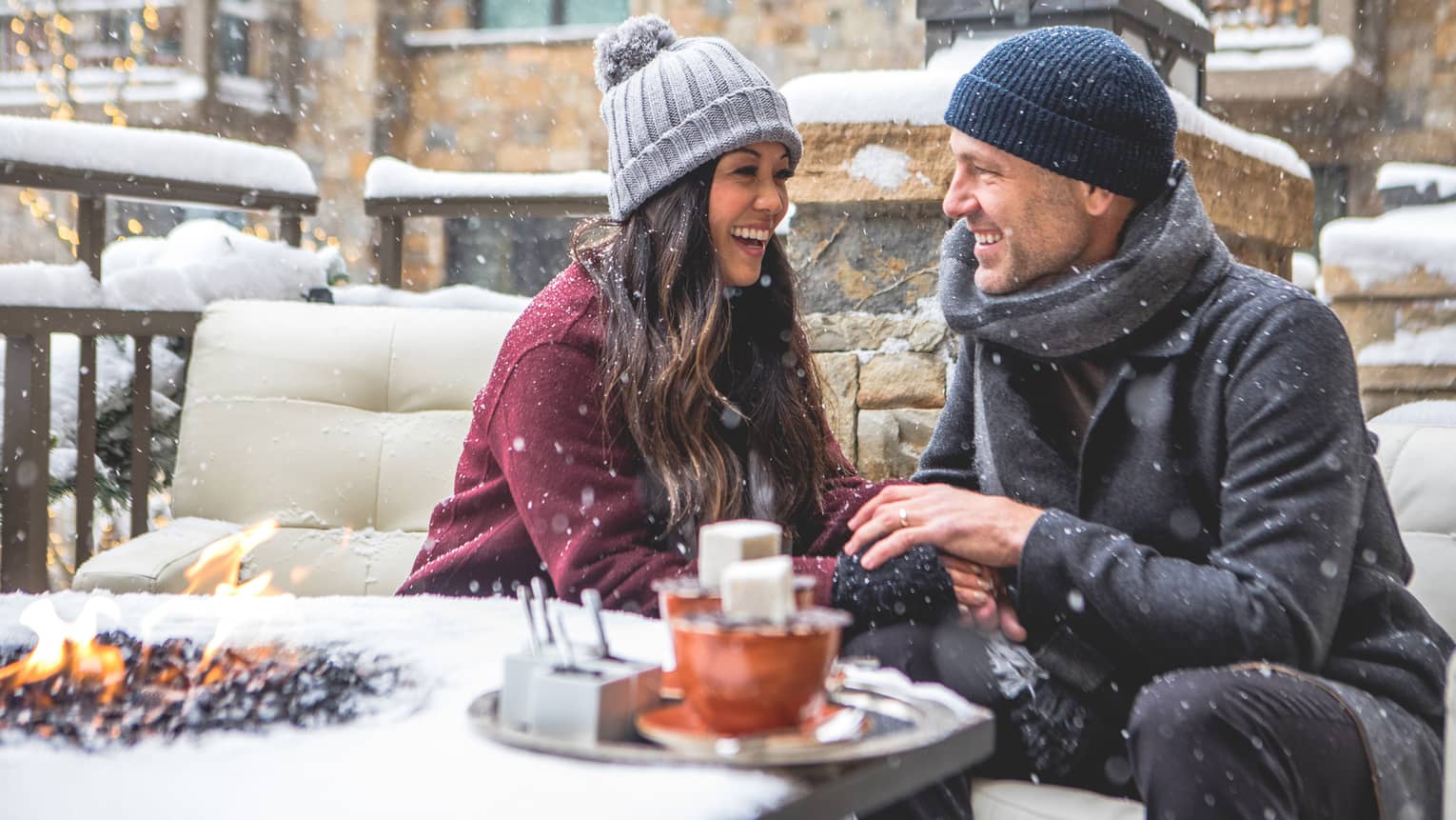 Smiling couple wearing winter clothes sit by hot chocolate tray, modern bonfire fireplace