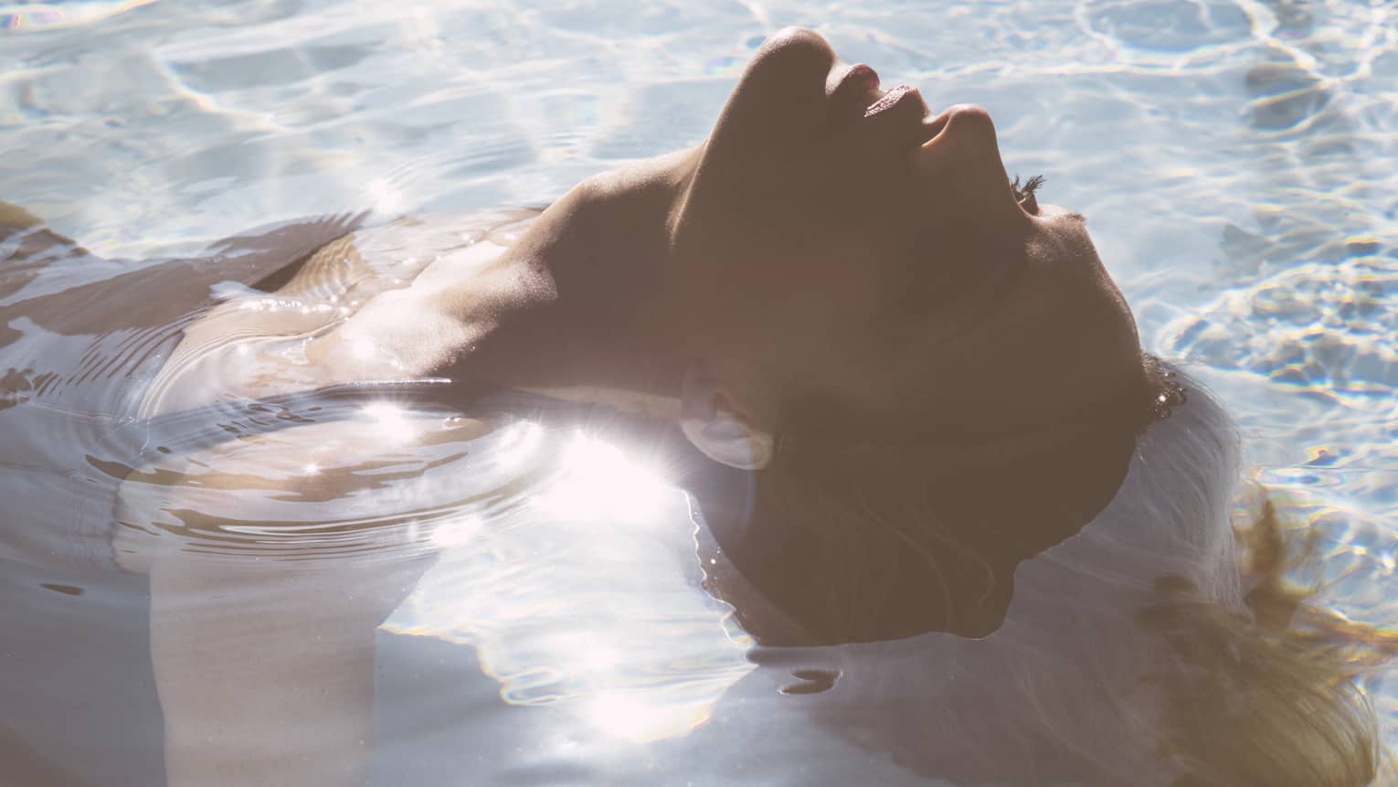 Close-up of woman's head as she floats on her back in pool, sunlight reflecting, her hair flowing in water 