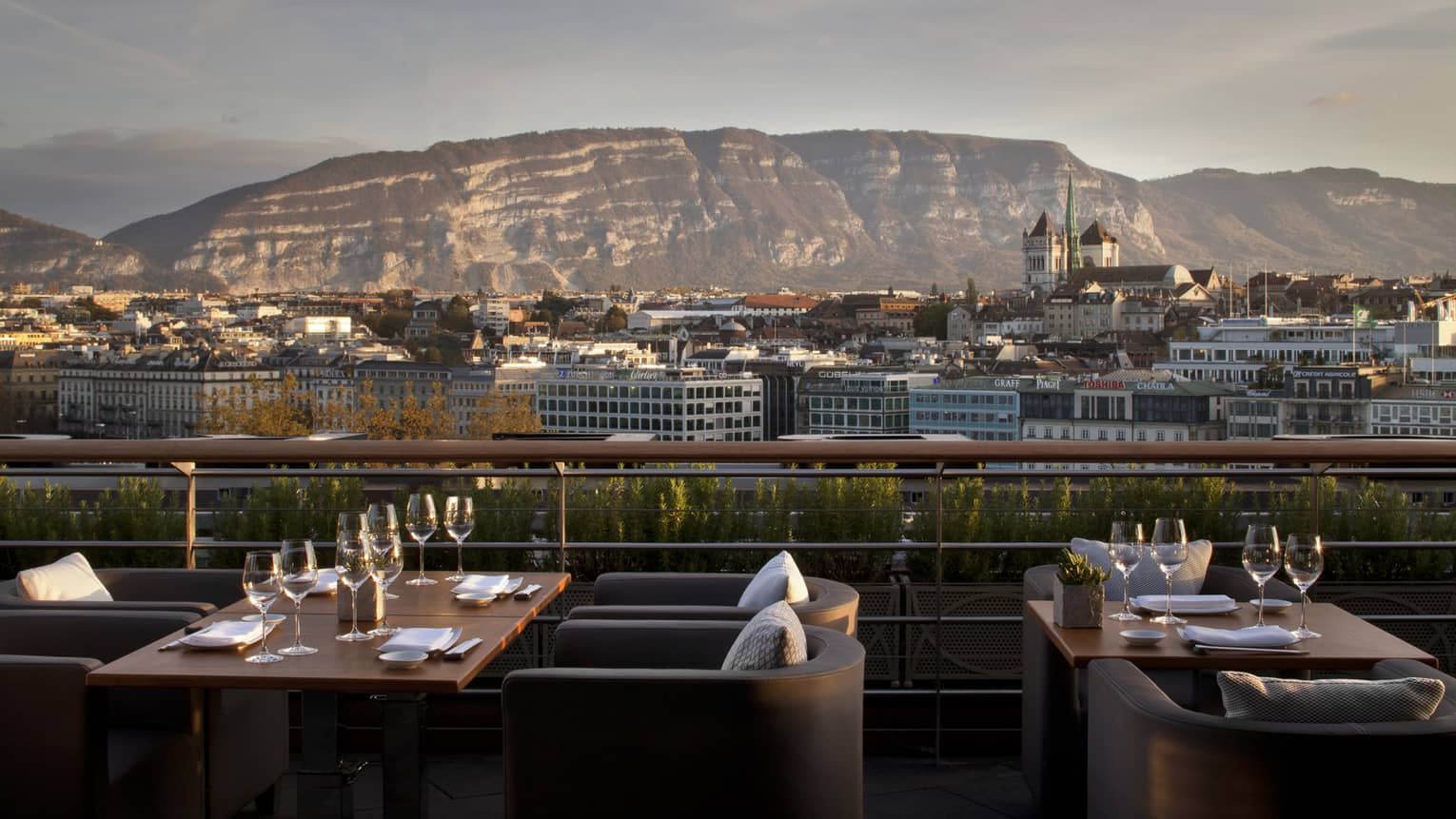 Outdoor patio with armchairs, tables set with wine glasses, overlooking city and mountain on sunny day