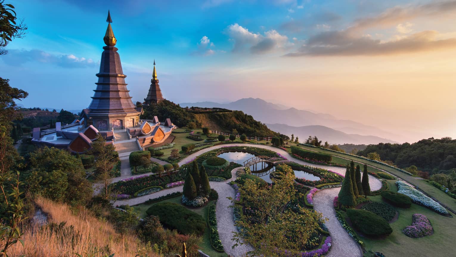 Two domed chedi monuments flanked by wending paths and manicured foliage atop a mountain, rose and gold sky and hills beyond.