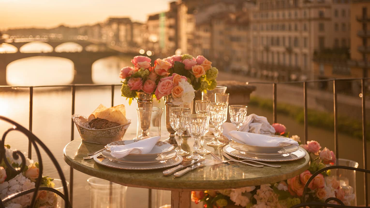 A romantic table for two is set with white porcelain plates, crystal glasses, pink roses on a balcony overlooking a bridge and historic building in Florence