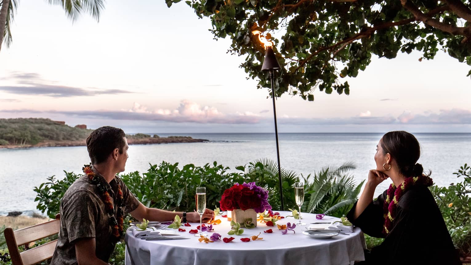 A couple wearing leis at a white table, and looking out on the ocean.