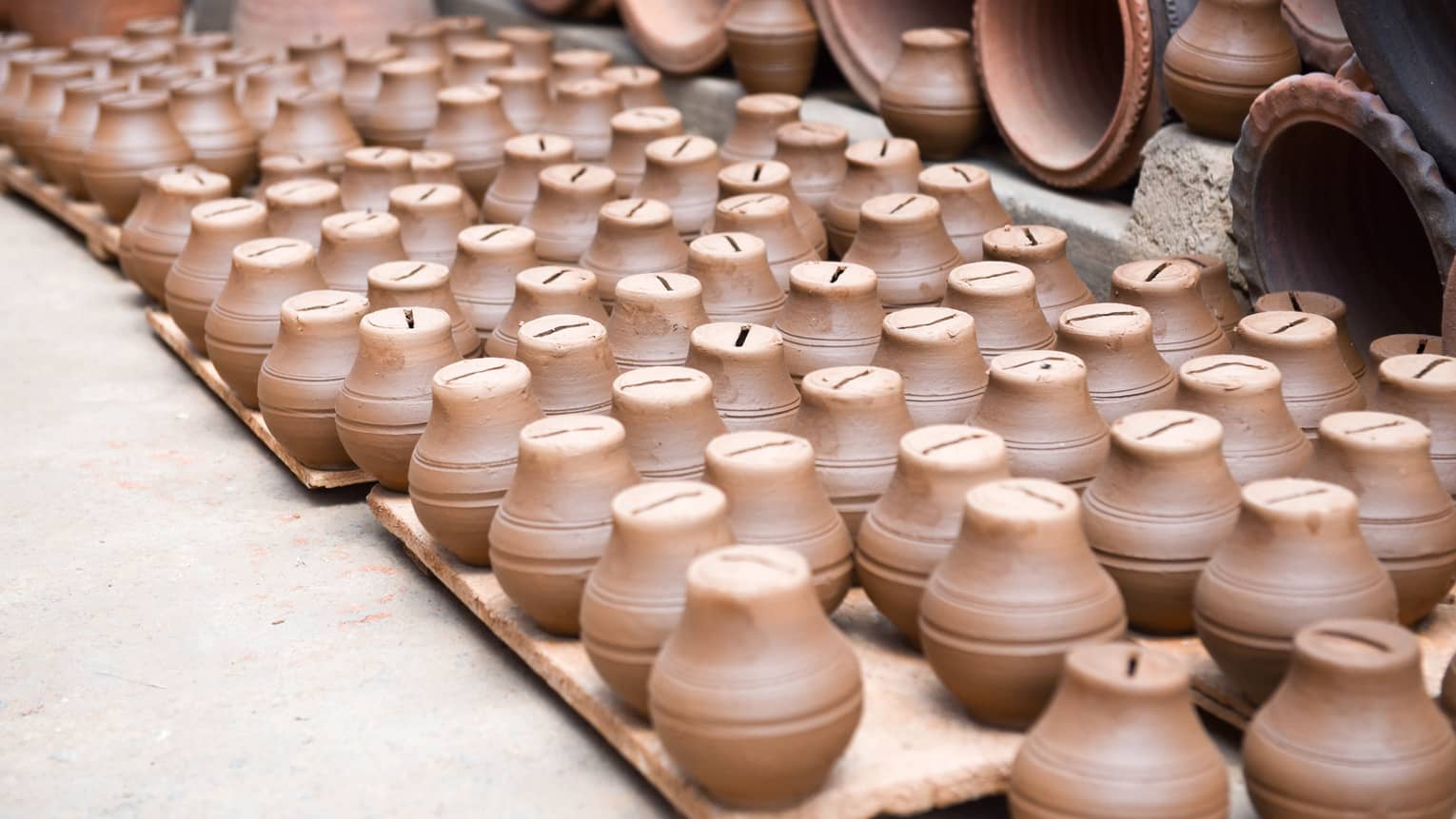 A collection of small, brown clay piggy banks neatly arranged on wooden pallets, surrounded by larger pottery items in the background, creating a display of handcrafted ceramics.