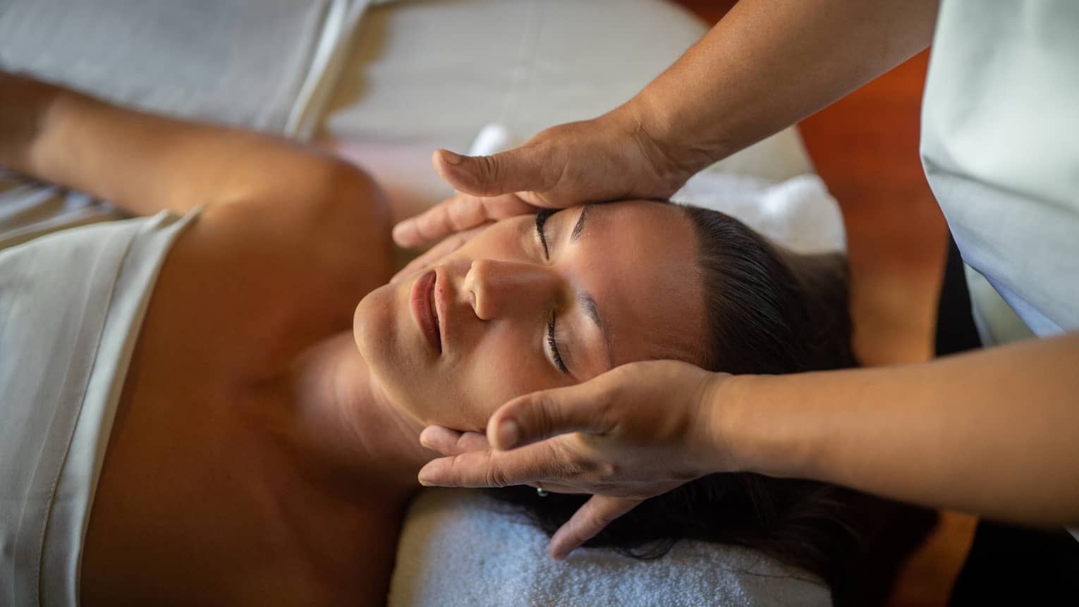 Woman lays on massage table with eyes closed as hands massage her cheeks and chin