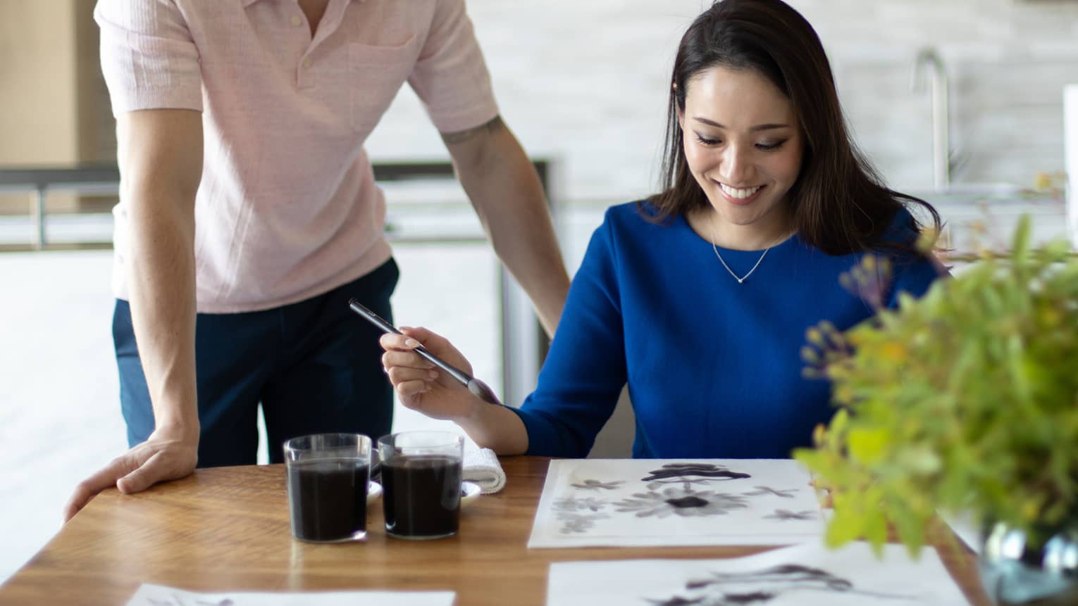 Smiling woman at table participates in Japanese ink wash painting while man stands over her should looking on
