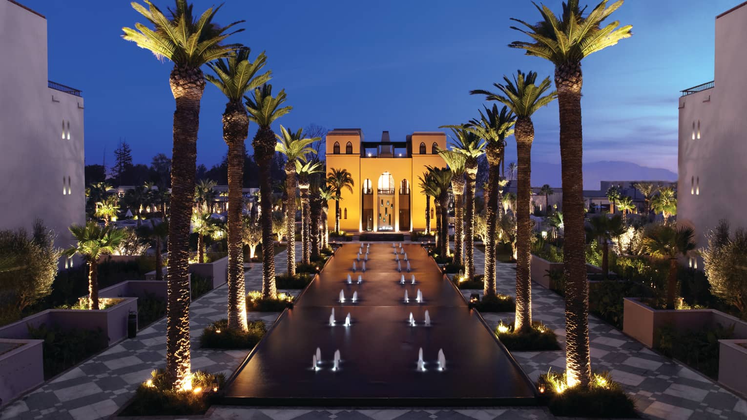 Fountains between rows of tall palm trees in front of illuminated Four Seasons Resort Marrakech hotel at night