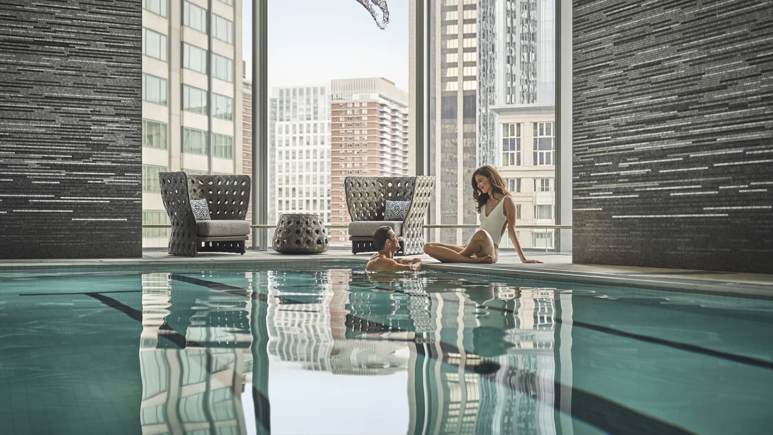 A couple embraces in a lap pool; behind them, floor-to-ceiling windows reveal an urban landscape.