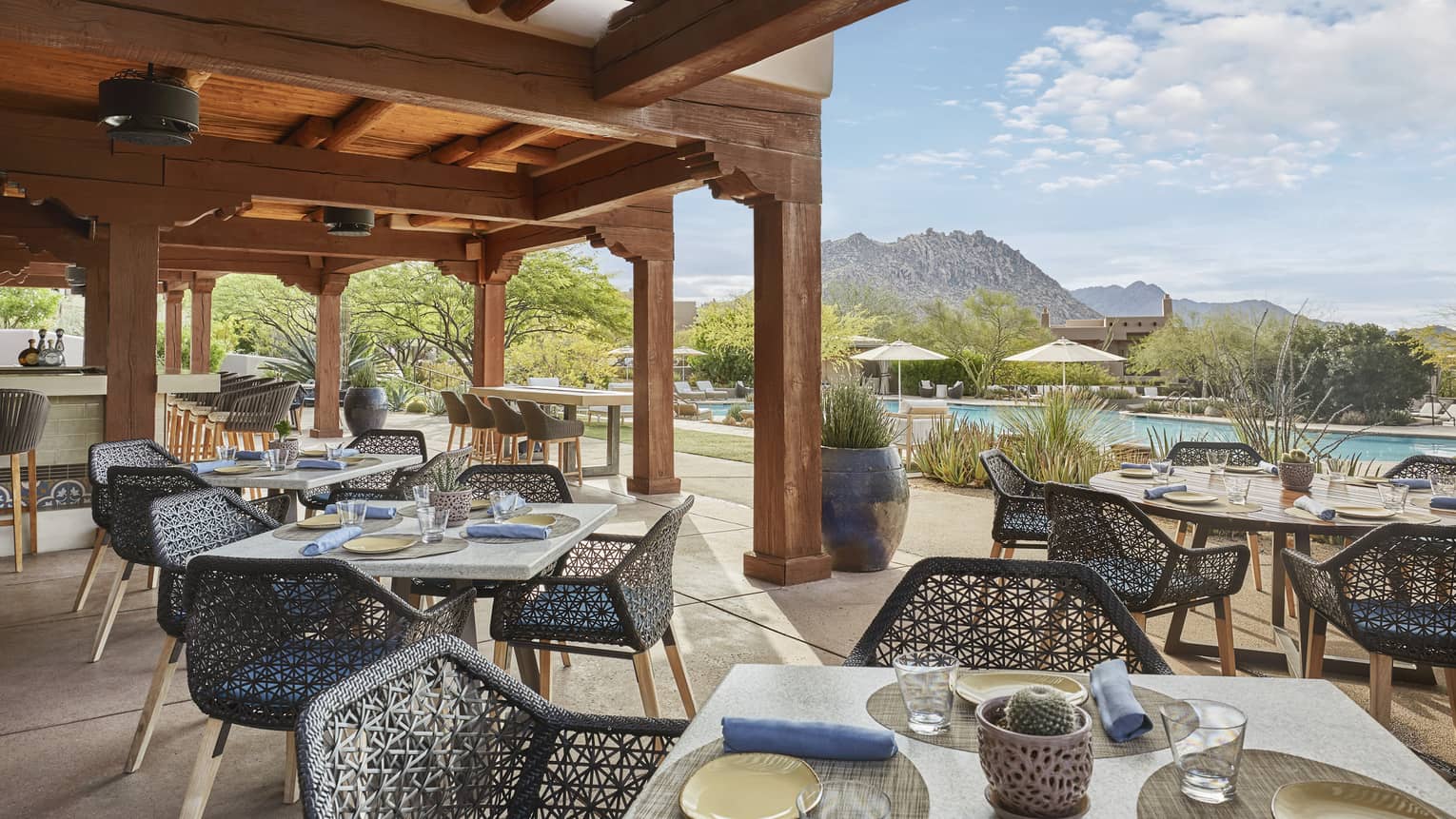 Outdoor restaurant patio with square tables, each with four dark blue wicker chairs, wooden pergola