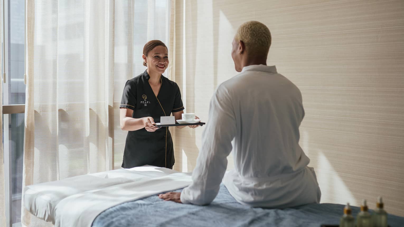 Spa attendant carrying a tea tray greets a man wearing a white spa robe who is sitting on a massage table
