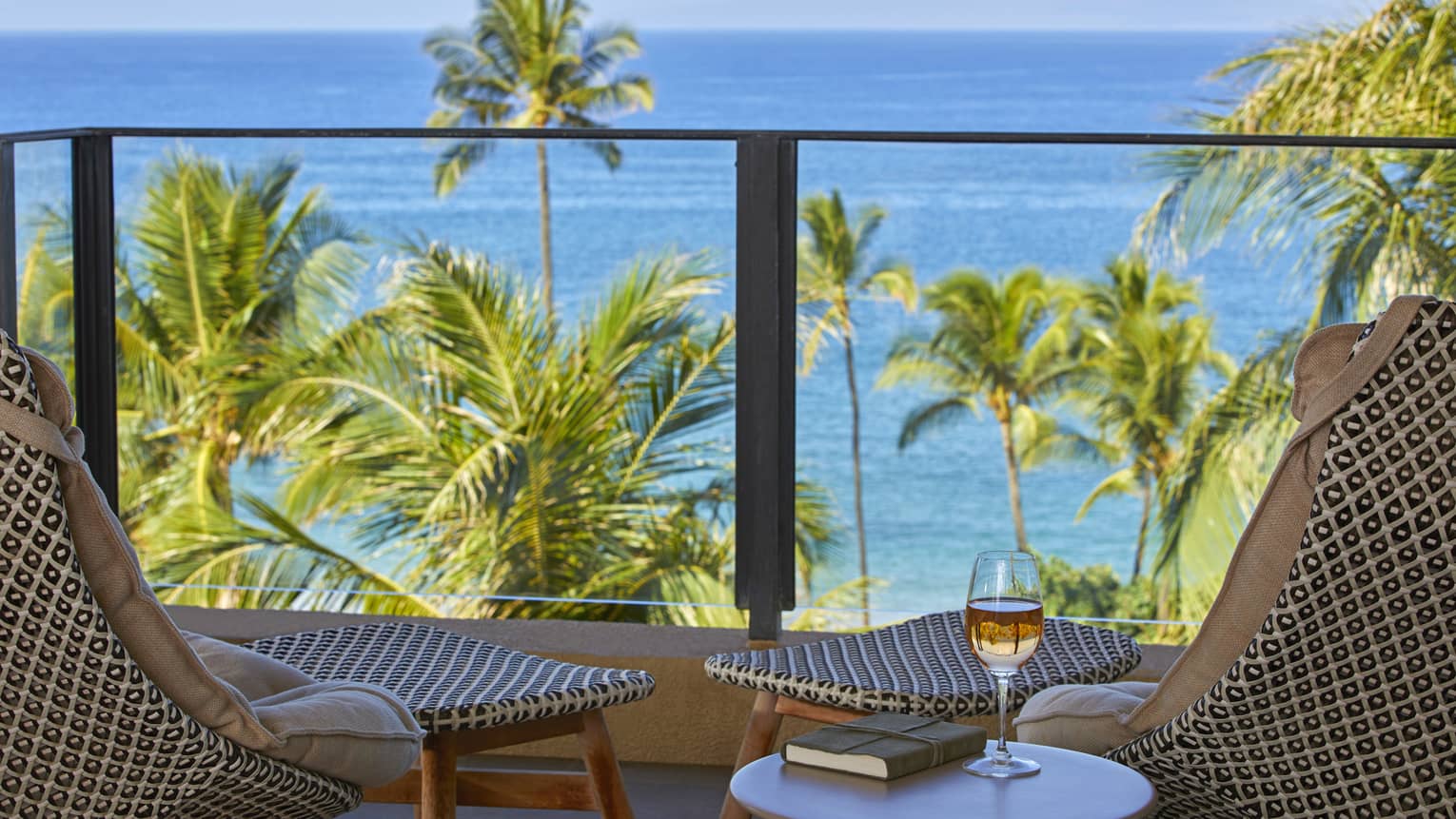 Balcony with rattan chairs, round table with book and glass of wine, overlooking palm trees and blue ocean