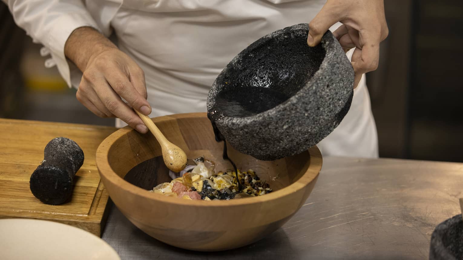 Chef pours black sauce from a stone bowl into a wood bowl of ingredients, a small wooden spoon in the other hand. 