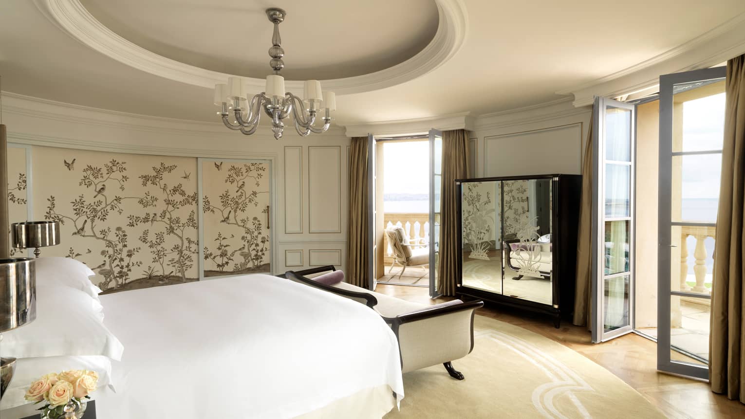 Round hotel room with recessed dome in ceiling, small chandelier, bed with white chaise, mirrored dresser, two patio doors