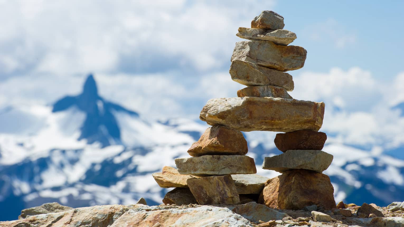 Close-up of an inukshuk atop a high rock shelf, against blue sky, white clouds and a majestic distant mountain.