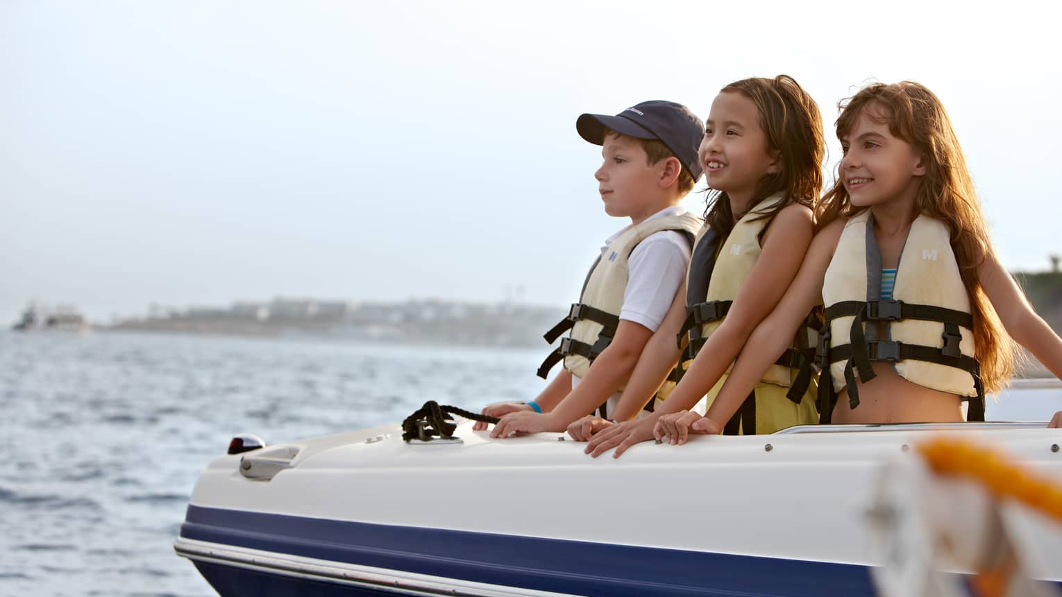 Three children in lifejackets in front of boat smile, look out on water 