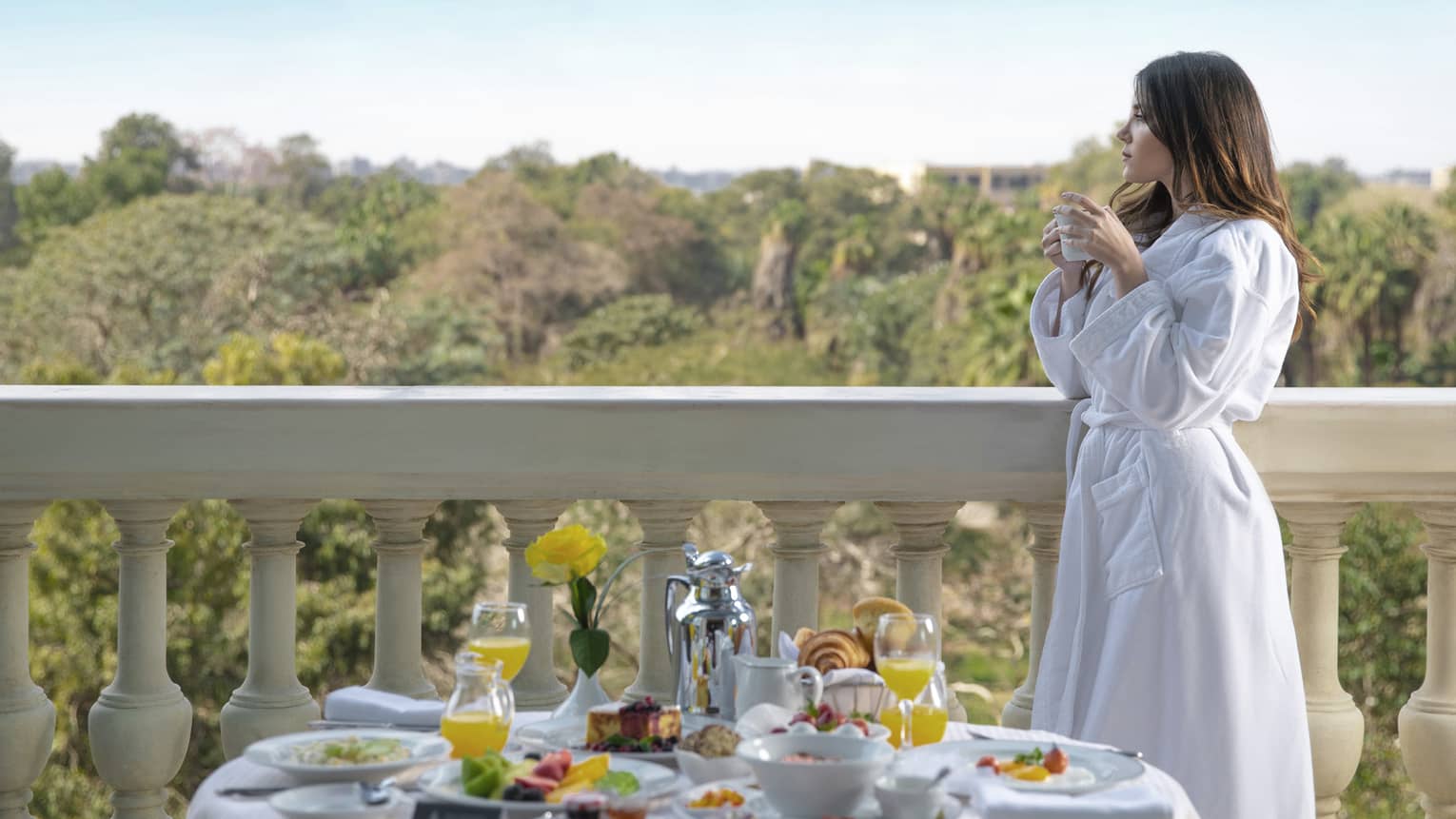 A woman in a white robe sips coffee while looking over a balcony, a variety of food on a table in front of her