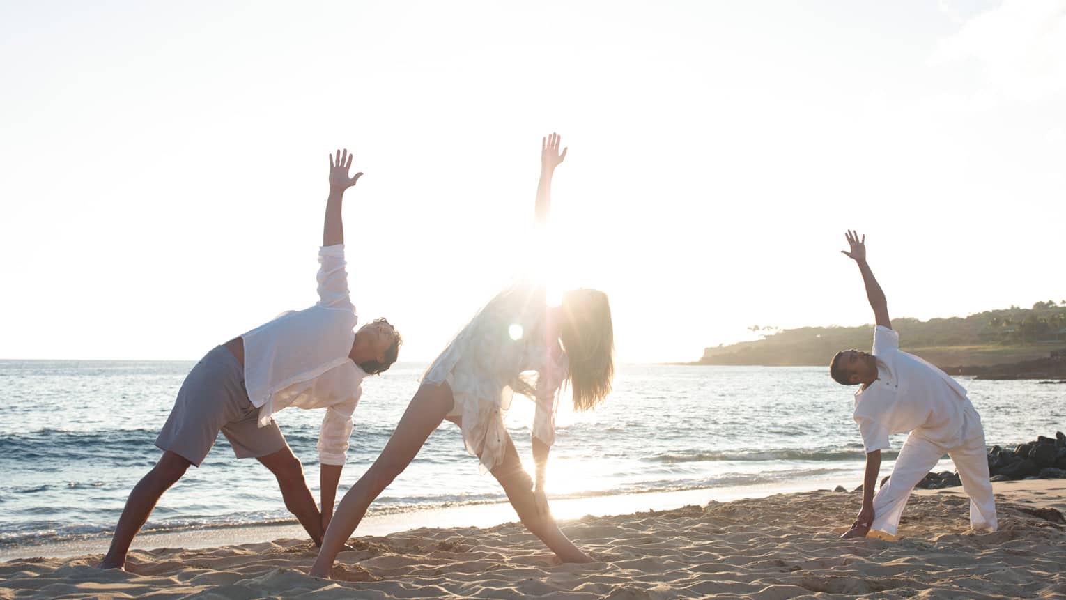 Three people stretch in yoga pose on sand beach at sunrise