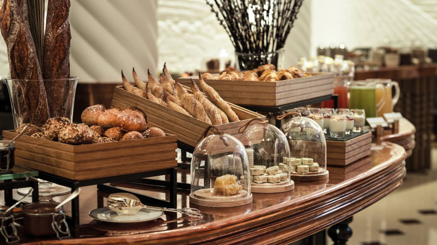 A decadent array of artisan breads and other baked goods in wooden crates and glass-covered dishes on a large wooden table.