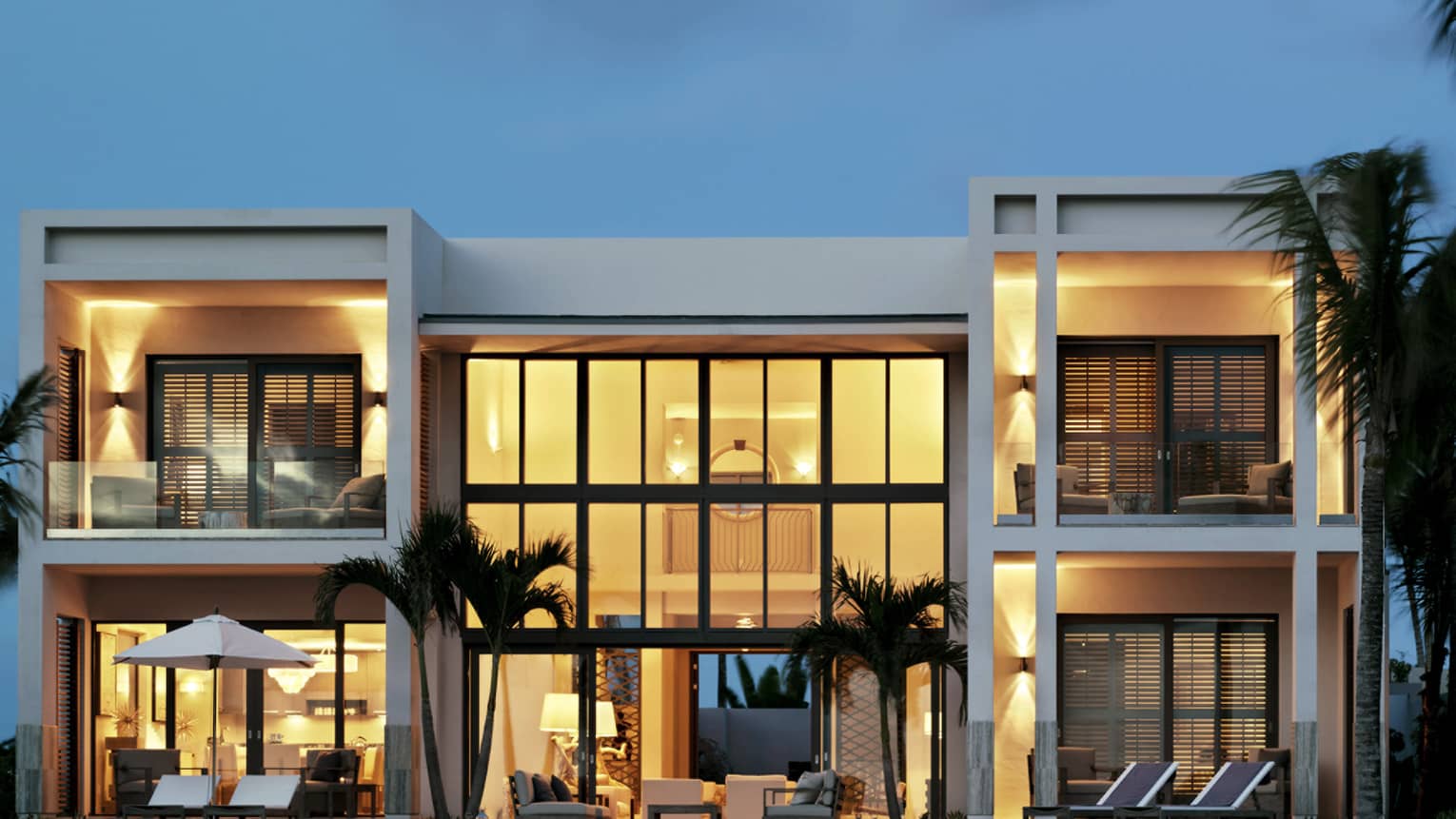 Exterior view of five-bedroom, white beachfront villa at dusk, two storeys with floor-to-ceiling illuminated windows