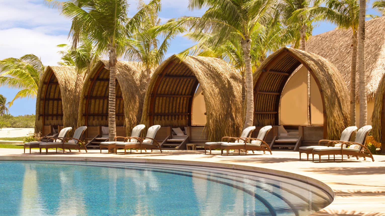 Four poolside pavilions with thatched roofs behind white lounge chairs, steps leading into swimming pool