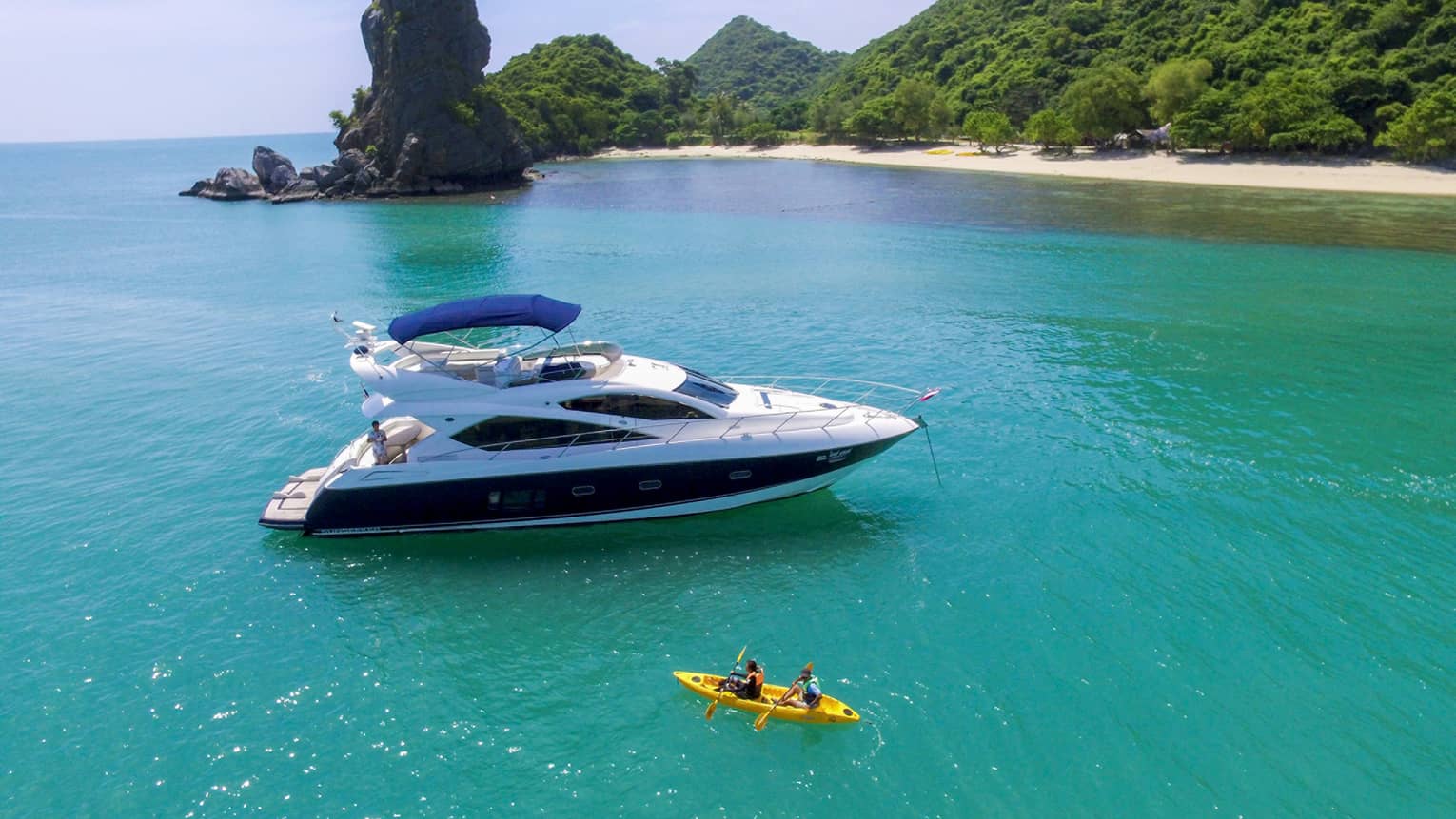 Two people in yellow kayak in front of large white yacht by island in blue ocean