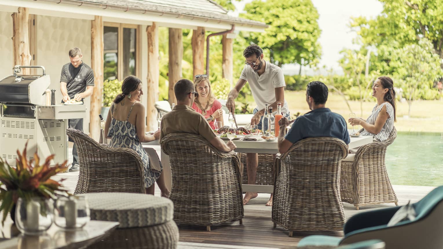 Five people seated poolside with staff member service and chef at grill