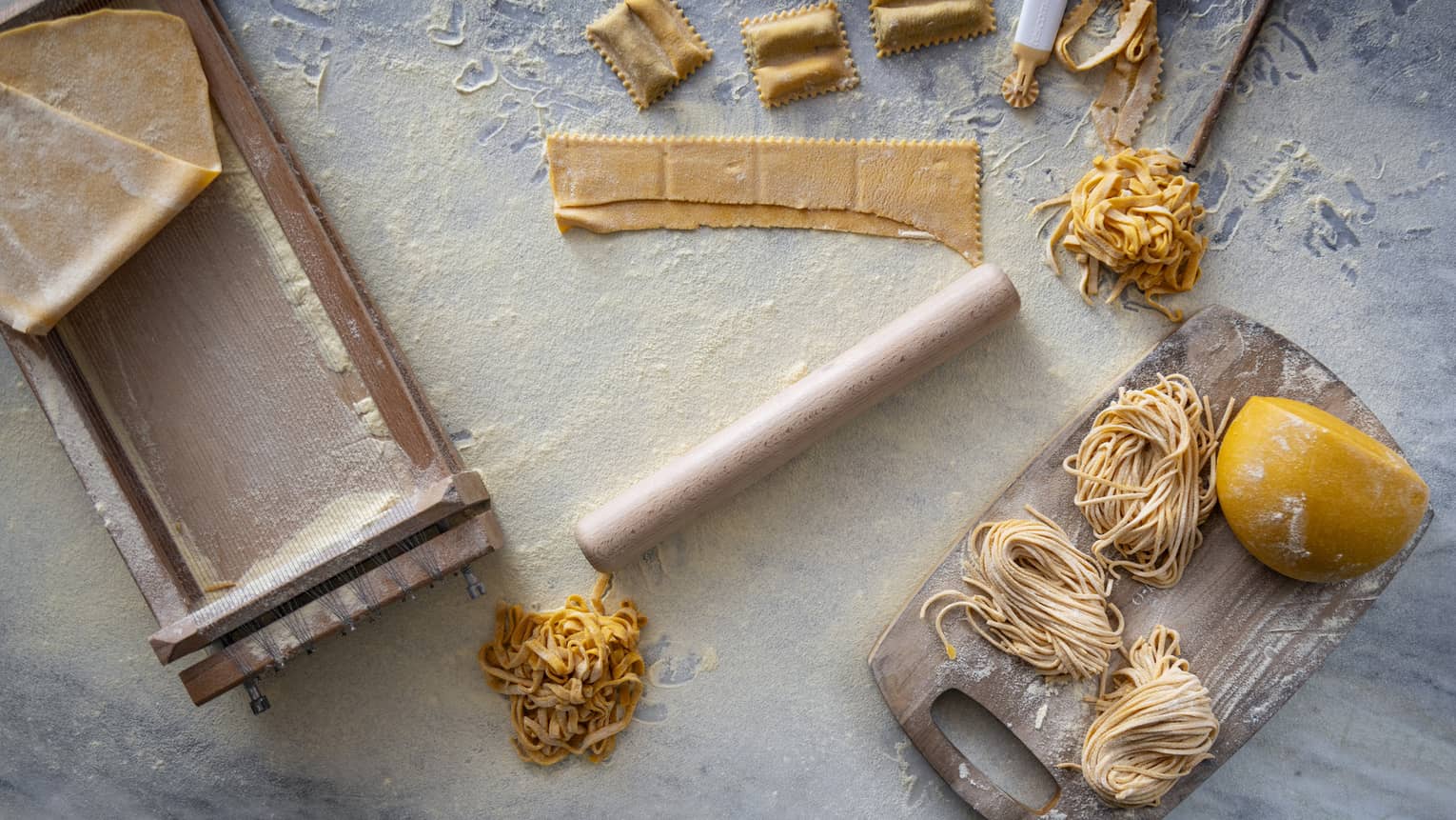 Marble slab covered in flour with a wooden rolling pin, pasta stretcher and cutting board topped with pasta in various states of completion