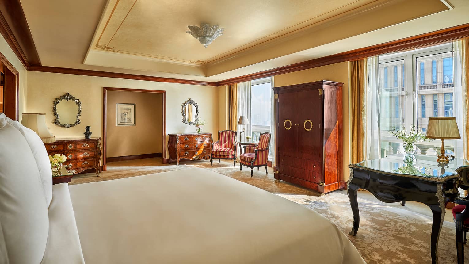 Hotel room bed across from two sunny floor-to-ceiling glass doors, antique-style wood dressers, armchairs 