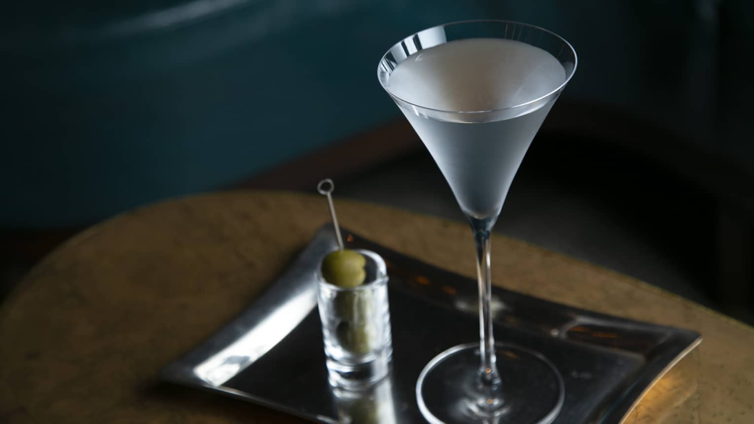 Signature cocktail in stemmed glass on silver tray, skewer of olives