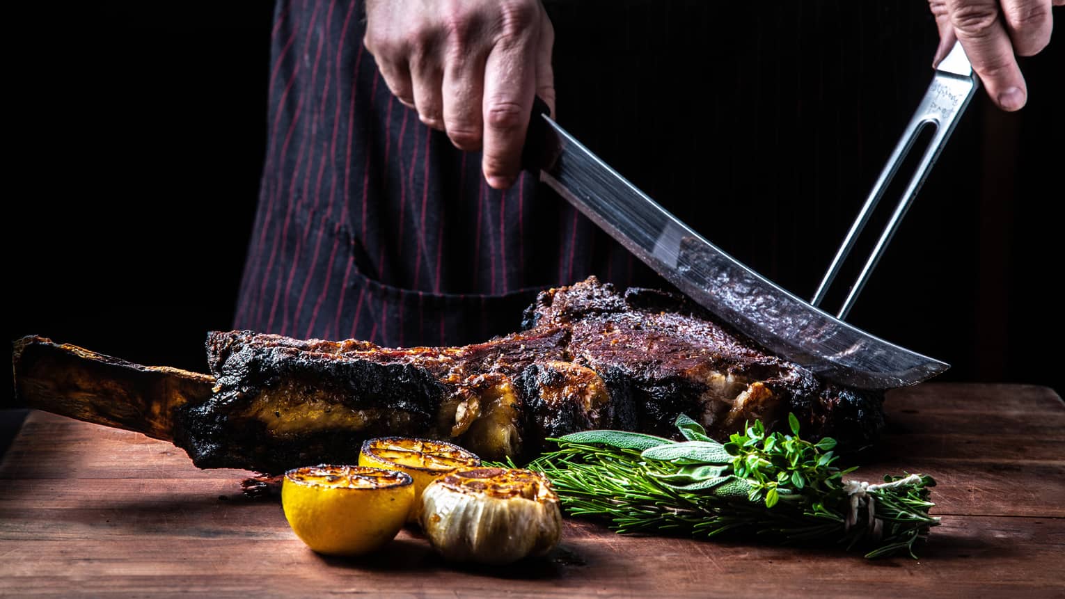 Close-up of chef slicing large rack of grilled meat, vegetables with butcher knives