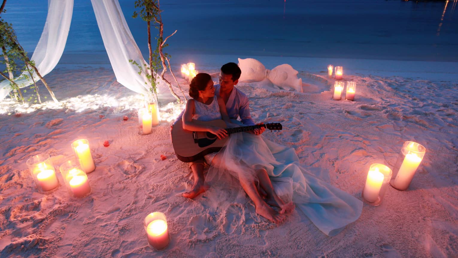 Man and woman hold guitar on sand beach surrounded by candles at night