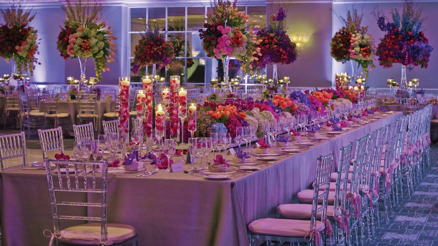 Large tropical flower-filled wedding banquet table in candlelit ballroom with purple lights