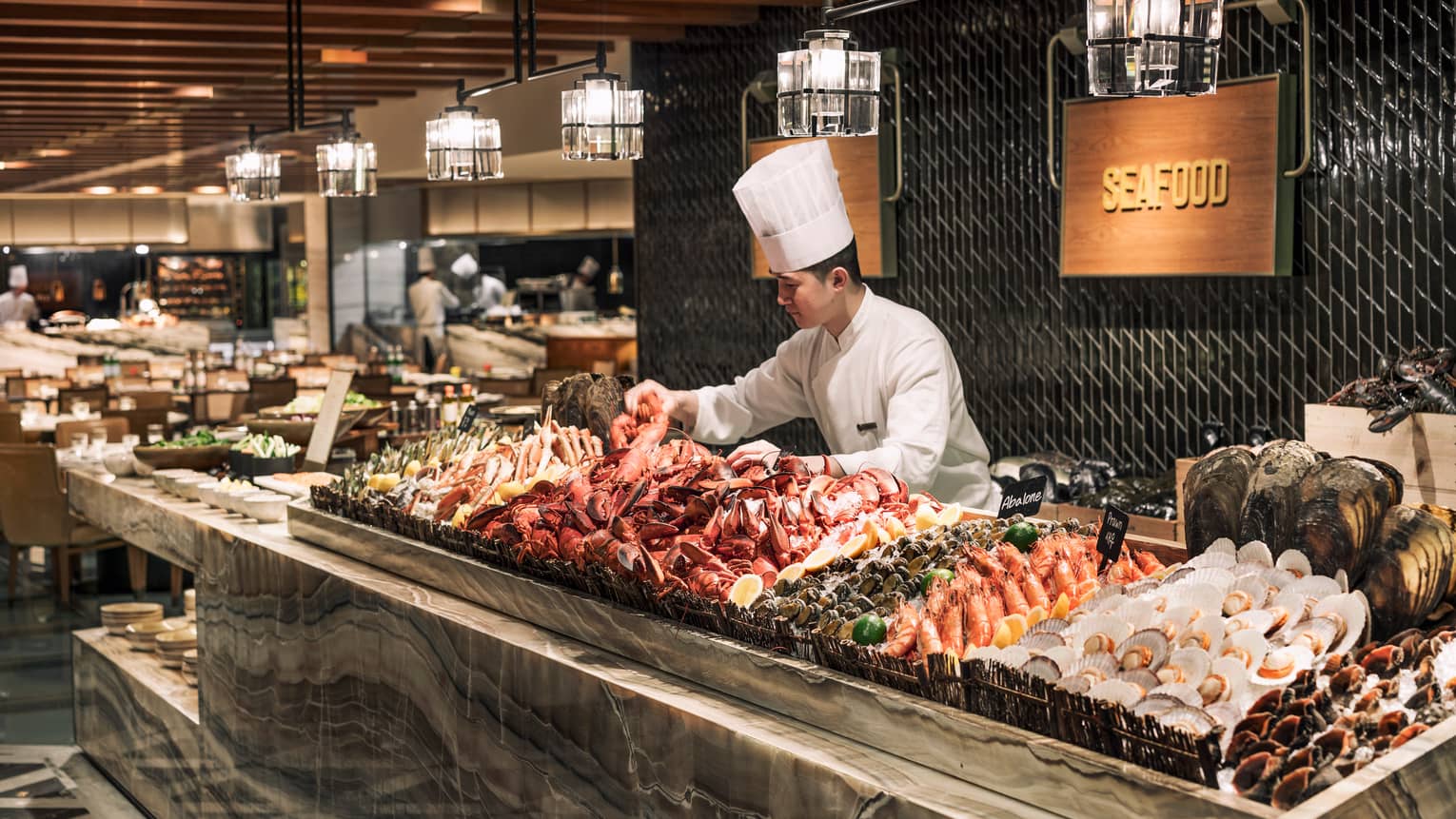 Chef wearing tall white hat arranges fresh lobster, seafood on ice at buffet The Market Kitchen buffet station