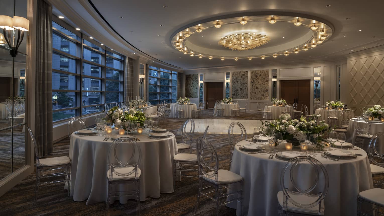 A dimly lit large meeting room with round tables covered in flowers and large windows.