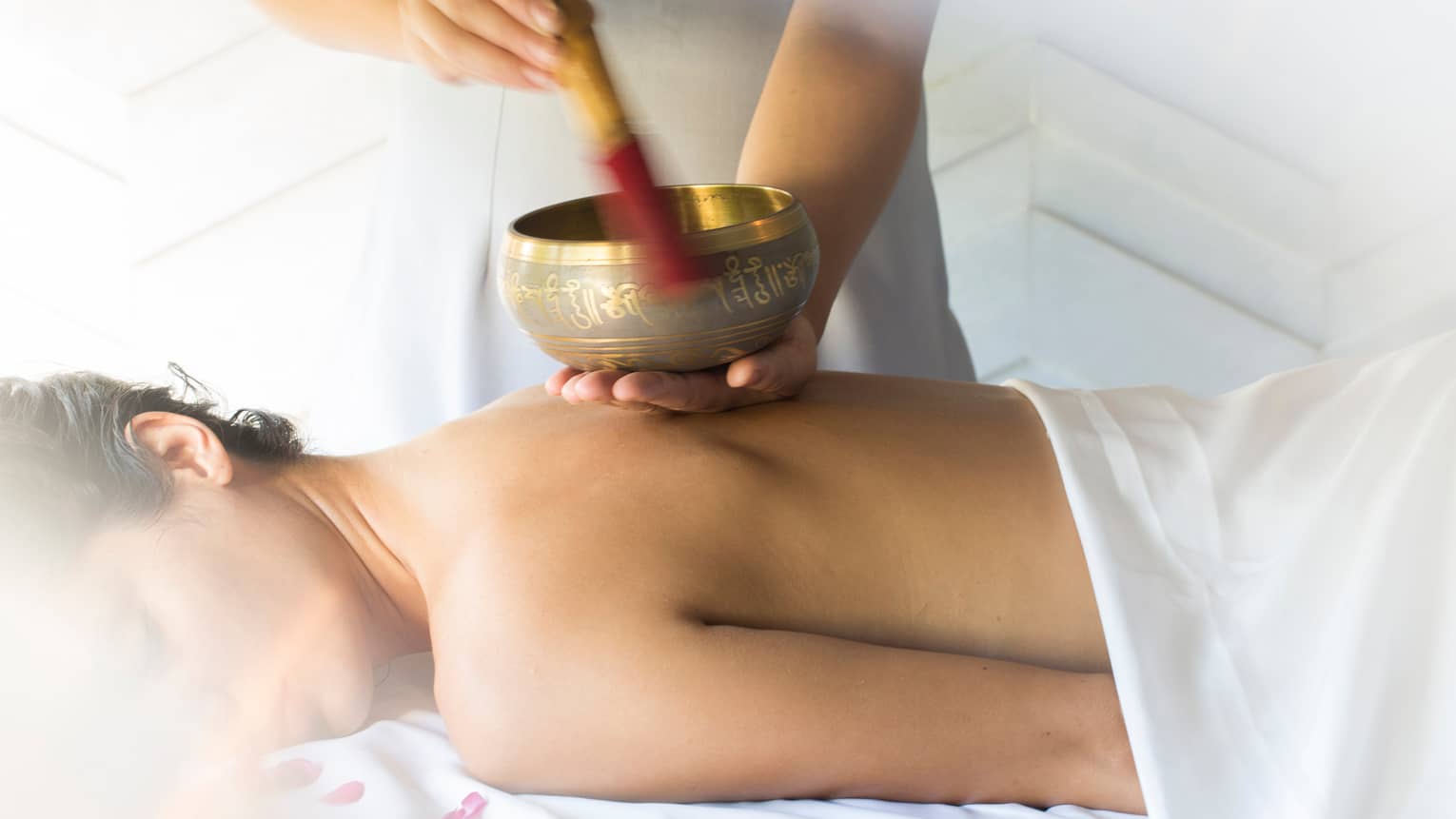 Hands play brass singing bowl over bare back of woman lying on massage table