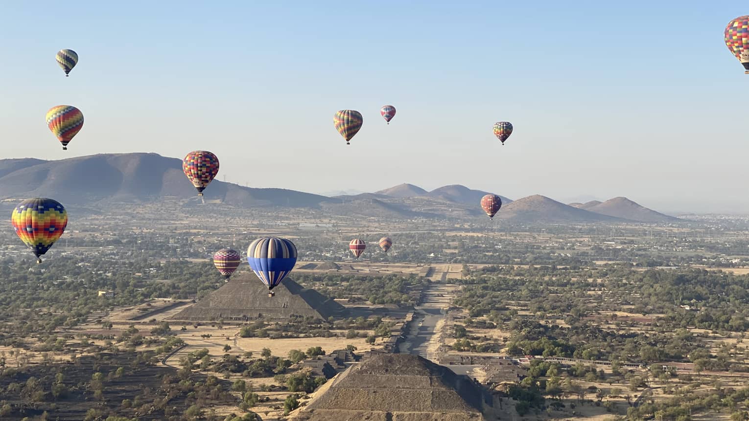 Hot-air balloons floating over old temples in a dessert covered in shrubs.