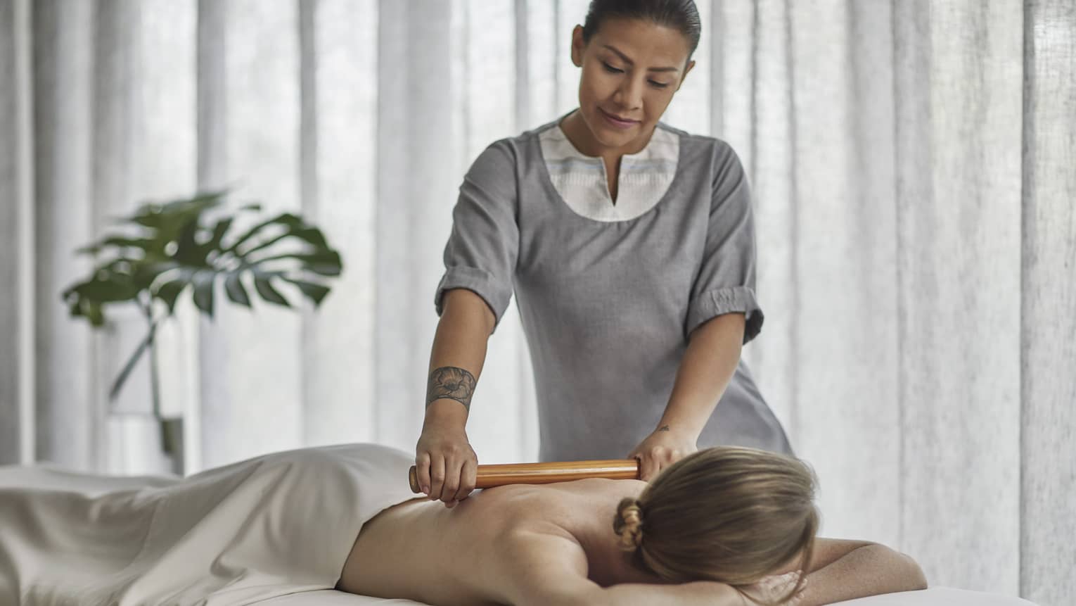 Spa therapist wearing a grey tunic rolls a wooden roller on the bare back of a guest who is laying on a massage table 