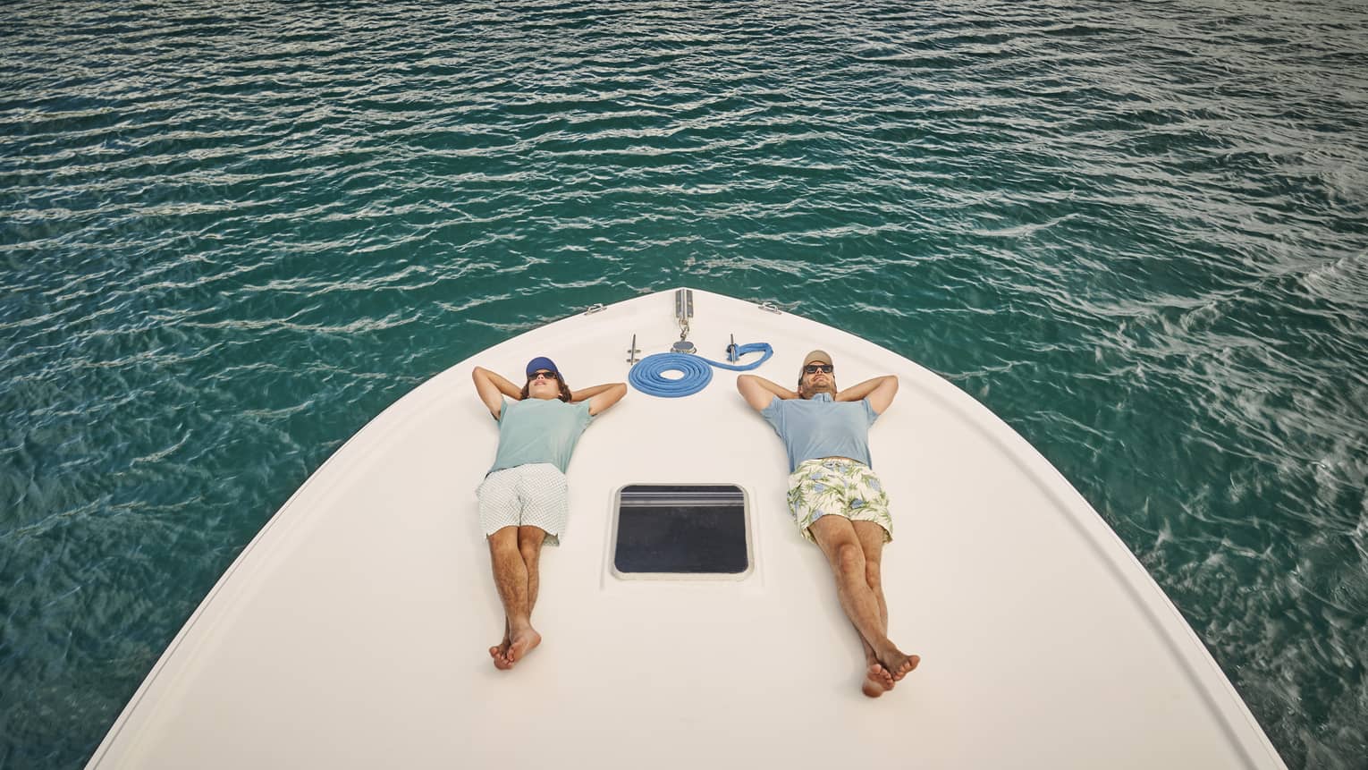Resting on a boat’s bow over gently rippling water, two passengers lie on their backs, legs crossed, hands behind heads.