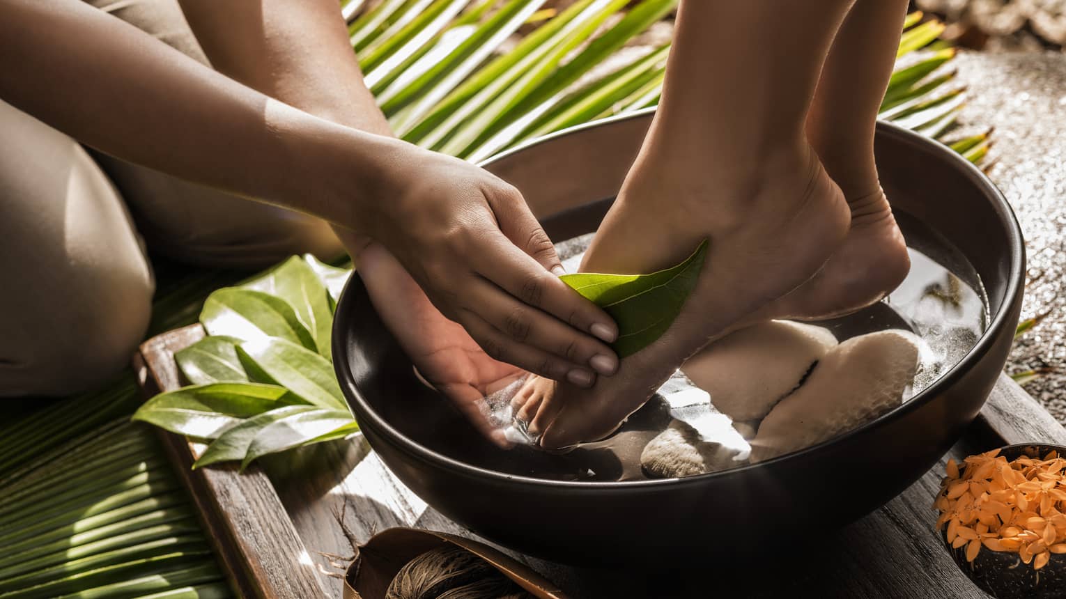 Close-up of woman's feet in round wood bowl as hands run tropical leaf over foot