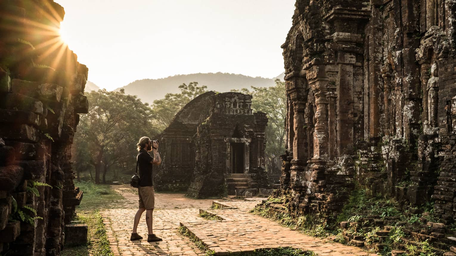 Tourist captures serene beauty of ancient Hindu temple ruins adorned with moss and greenery, sun streaming through the mist.