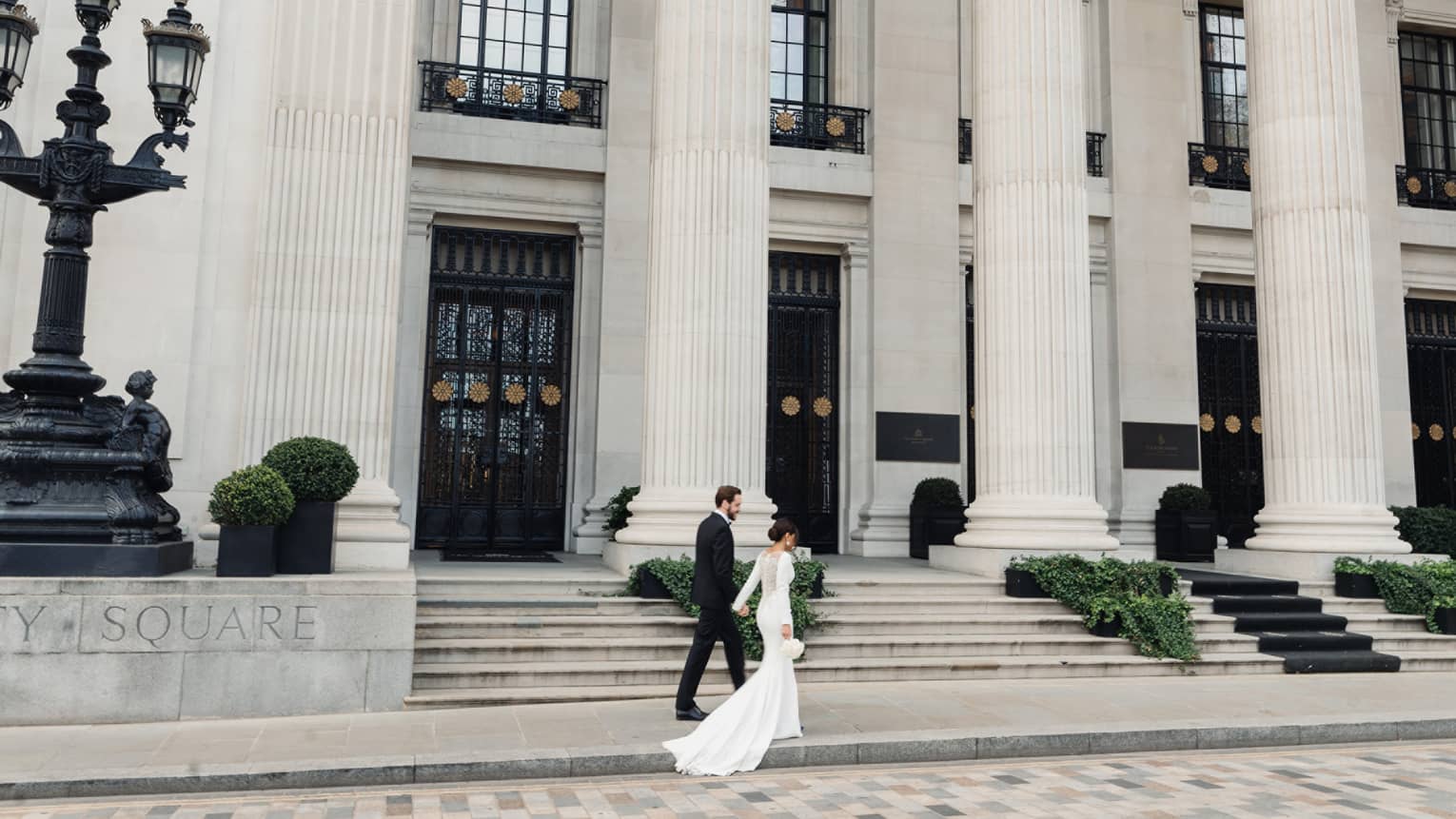 A bride and groom walking along a stone pathed street next to a large stone building with columns.