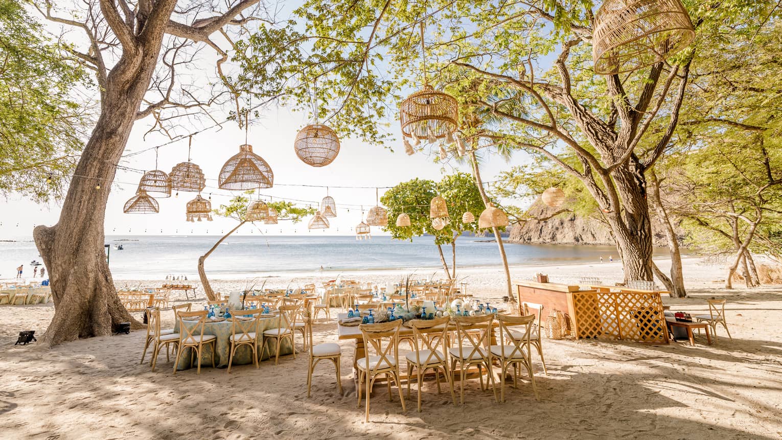 A group of tables with eight seats each are set on the beach surrounded by trees with a variety of straw light fixtures hanging from lines strung between the branches
