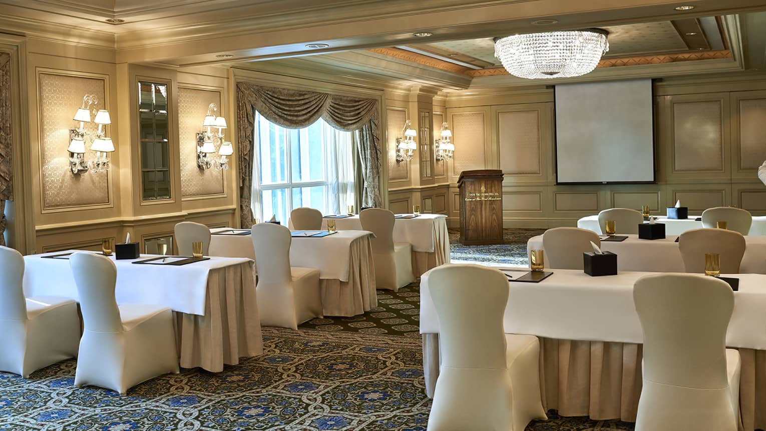 Rows of small meeting tables, chairs with white linens facing conference podium in event room