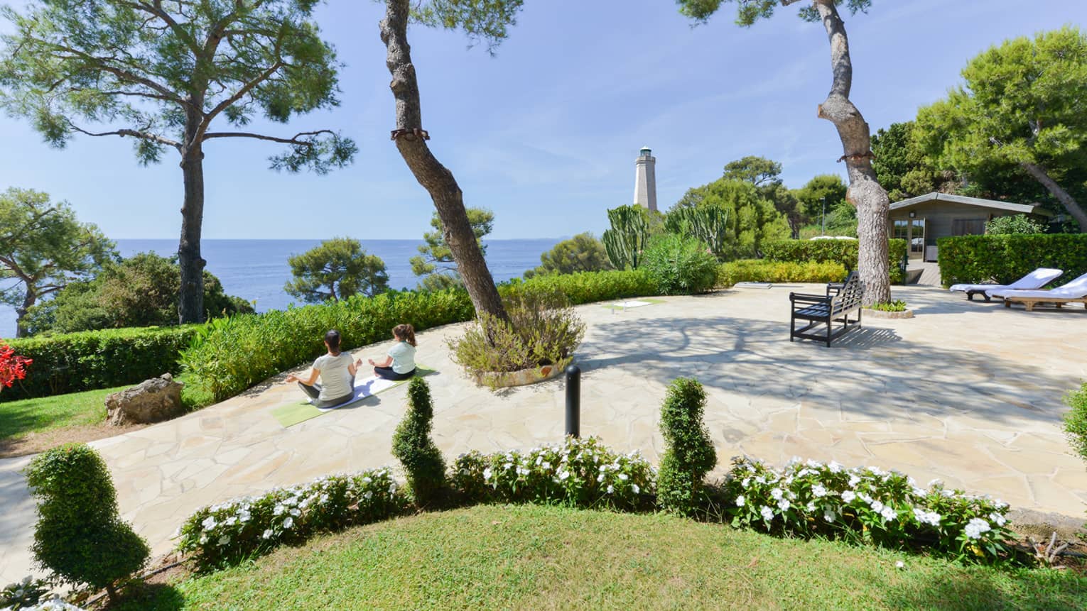 Rear view of two people in a seated yoga pose on curved patio with lush greenery, sea and lighthouse view